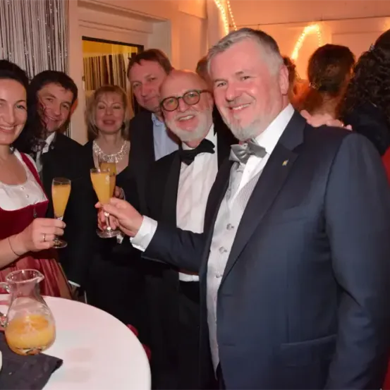 Eine Gruppe von Menschen in formeller Kleidung, wahrscheinlich auf einer Party, stoßen mit Orangengetränken an. Eine Frau in einem roten Kleid und ein Mann in einem Anzug mit einer Fliege sind vorne.