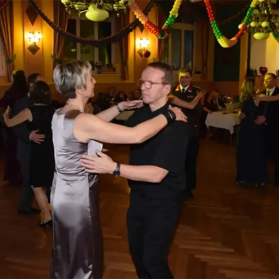 Ein Mann und eine Frau tanzen zusammen in einem Ballsaal. Sie sind von anderen Tänzern umgeben. Der Raum ist mit bunten Girlanden geschmückt.