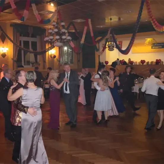 Ein mit bunten Dekorationen und herzförmigen Luftballons geschmückter Ballsaal voller tanzender Menschen. Einige Paare tanzen eng, während andere im Hintergrund stehen.