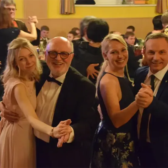 Mehrere Menschen in formeller Kleidung tanzen bei einer gesellschaftlichen Veranstaltung. Ein Mann und eine Frau tanzen zusammen, und ein weiterer Mann und eine Frau tanzen in der Nähe.