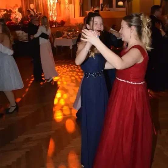 Zwei Frauen tanzen auf einer Tanzfläche bei einer Veranstaltung, eine in einem roten Kleid und die andere in einem blauen Kleid. Hinter ihnen tanzen oder stehen andere Menschen, mit einer Bühne im Hintergrund.