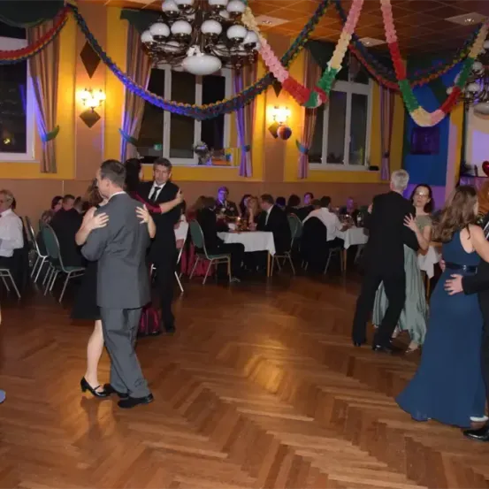 Mehrere Paare tanzen auf einem Holzboden mit einer Kronleuchter. Eine Menge beobachtet von Tischen. Der Raum ist mit bunten Wimpelketten und Luftballons dekoriert.