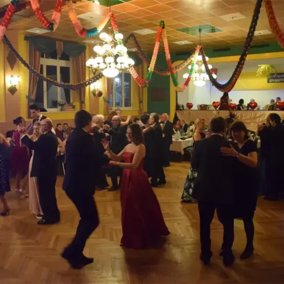 Mehrere Menschen tanzen in einem dekorierten Raum mit Kronleuchtern und einem Banner mit der Aufschrift 'Volksgarten Gloggnitzer'. Einige Leute sitzen an einem Tisch.