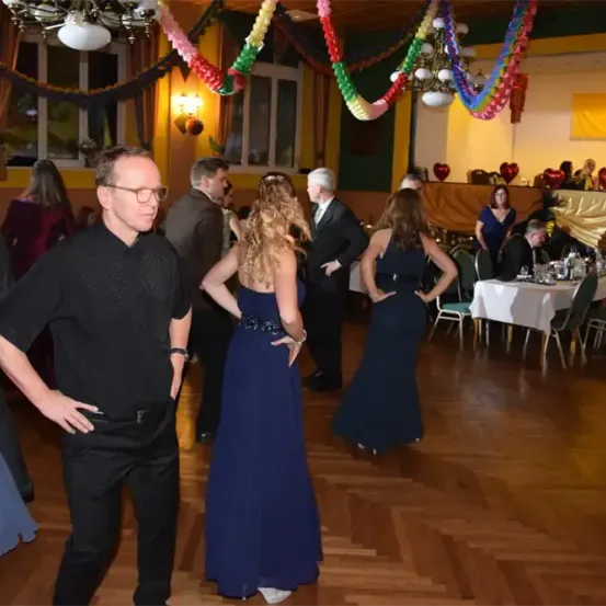 Eine Gruppe von Menschen versammelt sich in einem Raum mit Holzboden. Zwei Frauen tanzen, während andere in der Nähe stehen. Es gibt Stühle und Tische mit verschiedenen Gegenständen. Bunte Luftballons hängen von der Decke.