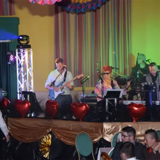 Eine Band tritt auf einer Bühne mit roten Herzdekorationen auf. Der Mann spielt Gitarre, während die Frau singt. Trommeln und eine Tastatur sind auf der Bühne. Das Publikum sieht der Aufführung zu.