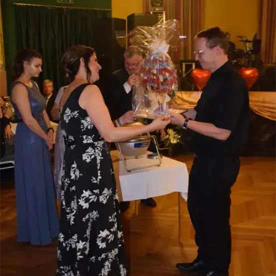 Ein Mann und eine Frau stehen und halten einen Geschenkkorb mit einem Blumenstrauß darin. Eine weitere Frau in einem blauen Kleid steht in der Nähe und beobachtet. Dahinter sitzt ein Mann, mit weiteren Dekorationen im Hintergrund.