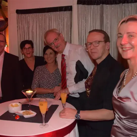 Sechs Personen, darunter vier Männer und zwei Frauen, versammeln sich um einen Tisch und posieren lächelnd für ein Foto. Sie halten Getränke in Gläsern, und auf dem Tisch befinden sich eine Schüssel, ein Teller mit Essen und eine Kerze. Hinter ihnen hängen zwei Vorhänge.