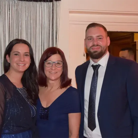 Drei Menschen posieren für ein Bild bei einer Veranstaltung. Der Mann trägt Anzug und Krawatte, die Frauen Abendkleider. Sie stehen eng beieinander.