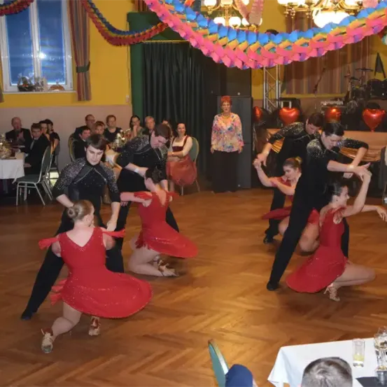 Eine Gruppe von Tänzern führt auf einer Tanzfläche in einem dekorierten Raum auf. Einige Tänzerinnen tragen rote Kleider, und der Raum hat eine gelbe Wand und einen Holzboden. Zuschauer sitzen um Tische herum.