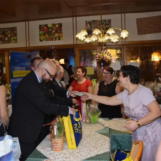 Ein Mann in einem Anzug überreicht einer Frau einen Geschenkbeutel bei einer Veranstaltung. Mehrere Personen stehen um einen Tisch mit Geschenken. Eine Hängelampe hängt von der Decke.