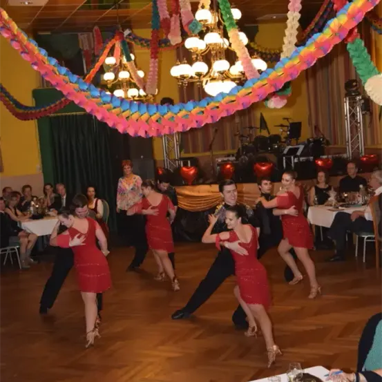 Eine Gruppe von Paaren in roten Kleidern tanzt auf einem dekorierten Tanzboden, mit einer Bühne, Musikinstrumenten und Zuschauern im Hintergrund.