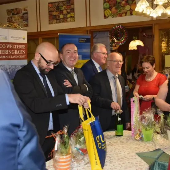 Eine Gruppe von Männern und Frauen in Anzügen und Kleidern lächelt und steht um einen Tisch mit Geschenktüten. Ein Mann hält eine gelbe Geschenktüte.