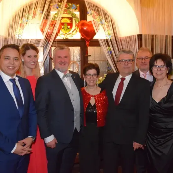 Eine Gruppe formell gekleideter Personen, darunter Männer und Frauen, steht zusammen für ein Foto. Eine Frau in einem roten Paillettenkleid steht in der Mitte. Hinter ihnen befindet sich ein farbenfrohes Buntglasfenster und ein roter Luftballon.