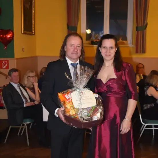 Ein Mann und eine Frau stehen zusammen und halten einen Geschenkkorb. Sie scheinen sich bei einer Veranstaltung zu befinden. Hinter ihnen sitzen mehrere Personen an Tischen.