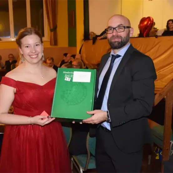 Ein Mann in Anzug und eine Frau in rotem Kleid halten ein grünes Buch. Sie lächeln und schauen in die Kamera. Dahinter eine Bühne mit Luftballons und Publikum.