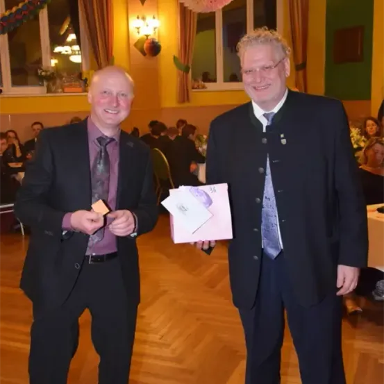 Zwei Männer stehen in einem Raum, lächeln und einer hält eine Karte, der andere einen rosa Umschlag. Sie sind beide in Anzügen und Krawatten gekleidet. Hinter ihnen sitzen Leute an Tischen und der Raum ist mit festlichen Dekorationen geschmückt.