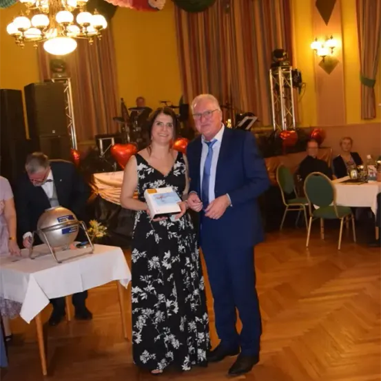 Ein Mann und eine Frau stehen zusammen auf einer Tanzfläche und halten eine Geschenkbox. Sie sind umgeben von einem dekorierten Raum mit Tischen, Stühlen und einer Band, die im Hintergrund aufgebaut ist.