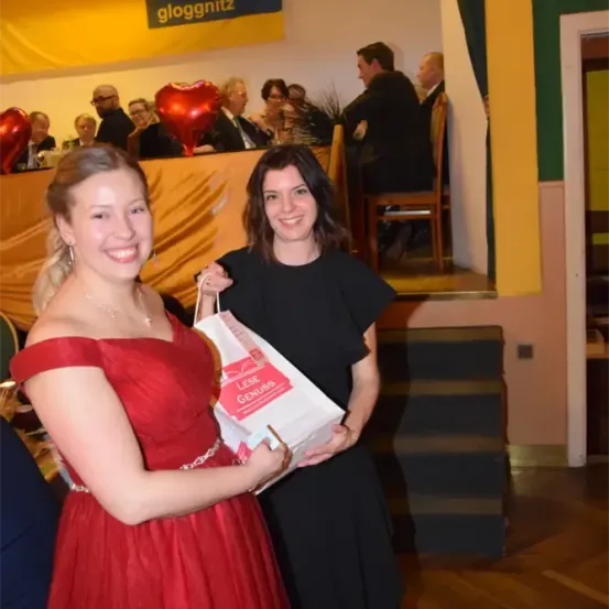 Zwei Frauen halten einen Geschenkbeutel. Eine Frau trägt ein rotes Kleid, die andere ein schwarzes Kleid. Hinter ihnen sitzen Menschen an Tischen, und ein Banner zeigt 'volkspartei gloggnitz'.
