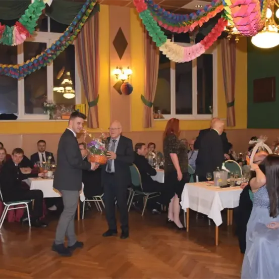 Eine formelle Veranstaltung findet in einem Saal statt. Zwei Männer präsentieren einer Frau einen Blumenstrauß. Mehrere Personen sitzen an Tischen. Der Saal ist mit bunten Bändern dekoriert und hat gelbe Wände.