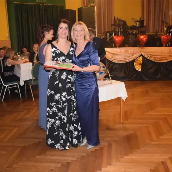 Zwei Frauen in Abendkleidern stehen auf einer Tanzfläche, eine hält ein Buch. Hinter ihnen steht ein Tisch mit einer herzförmigen Dekoration. Menschen sitzen an Tischen und eine Bühne mit Musikinstrumenten ist im Hintergrund.