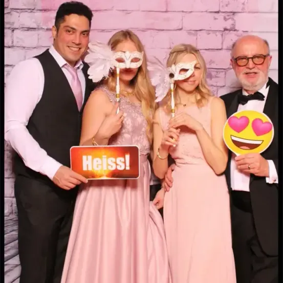 Drei Personen posieren für ein Foto mit rosa Masken und einem Schild mit der Aufschrift 'Heiss!' vor einer rosa Ziegelwand.