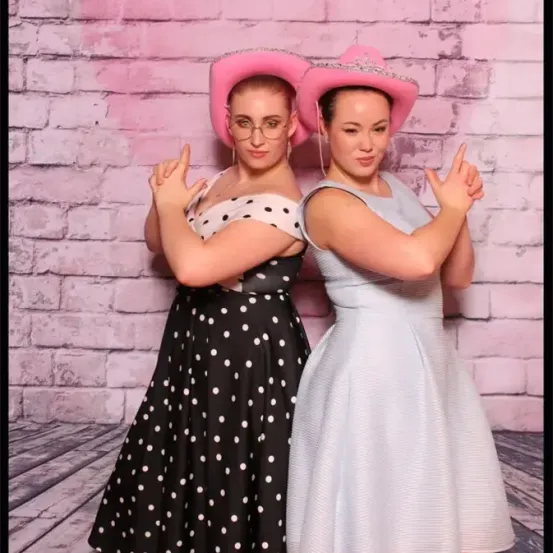 Zwei Frauen in Retro-Kleidung posieren für ein Foto vor einer rosa Ziegelwand. Eine trägt ein gepunktetes Kleid und die andere ein gestreiftes Kleid, beide mit Cowboy-Hüten.