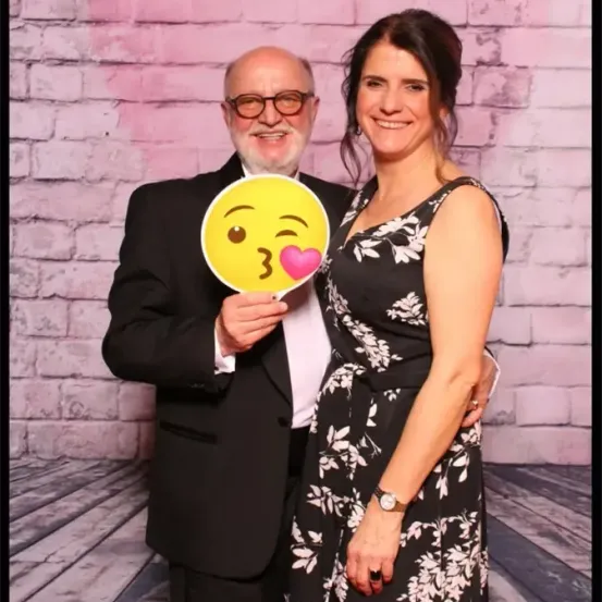 Ein älterer Mann und eine Frau stehen lächelnd nebeneinander und posieren für ein Foto vor einer rosafarbenen Ziegelwand. Der Mann hält einen gelben Aufkleber mit einem lächelnden Gesicht und einem Herz.