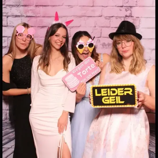 Vier Frauen stehen vor einer rosa Ziegelwand, tragen Brillen und Requisiten. Eine hält ein Schild mit der Aufschrift 'wegen der Torte' und eine andere hält ein Schild mit der Aufschrift 'LEIDER GEIL'.