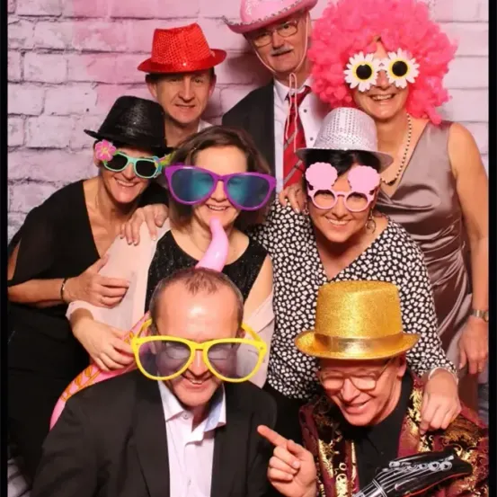 Eine Gruppe von Erwachsenen in lustigen Hüten und mit Sonnenbrillen posiert für ein Foto vor einer rosa Ziegelwand.