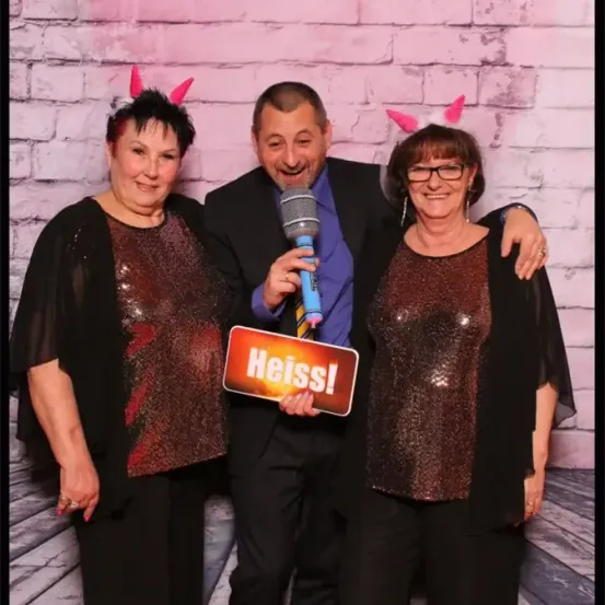Drei Personen posieren vor einer rosa Ziegelwand, alle mit festlichen Hasenohren. Der Mann hält ein Mikrofon, und ein Schild mit der Aufschrift 'Heiss!' wird in der Mitte gehalten.