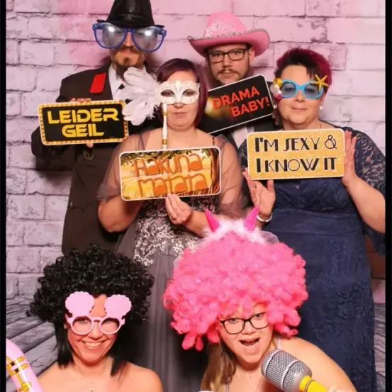 Eine Gruppe von Menschen in Kostümen und Brillen posiert für ein Foto. Sie halten Schilder mit den Aufschriften 'Leider Geil', 'Drama Baby!', 'Ich bin sexy und ich weiß es' und 'Aruna Mata'.