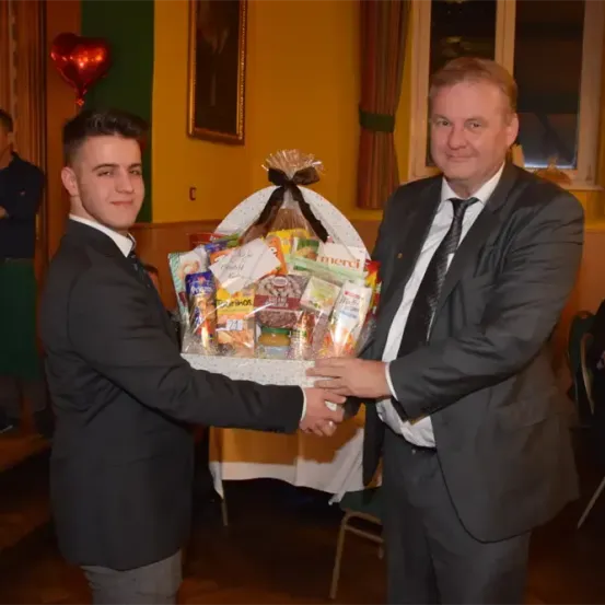 Zwei Männer in Anzügen tauschen einen Geschenkkorb in einem Raum mit gelber Wand und rotem Ballon aus.