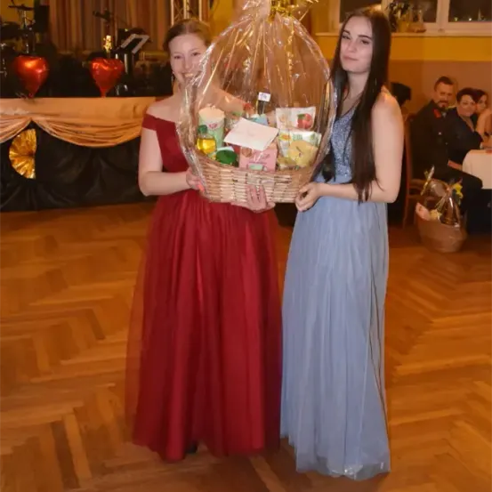 Zwei junge Frauen stehen auf einer Tanzfläche, eine hält einen Korb voller verschiedener Gegenstände, während die andere lächelt. Hinter ihnen steht ein Tisch mit einer herzförmigen Dekoration. Im Hintergrund ist ein verschwommenes Bild einer Person zu sehen, die an einem Tisch sitzt.