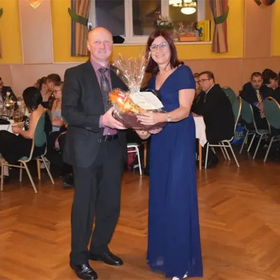 Ein Mann und eine Frau stehen zusammen und halten einen Geschenkkorb. Sie sind von sitzenden Gästen bei einem Bankett in einem Raum mit Holzboden umgeben.