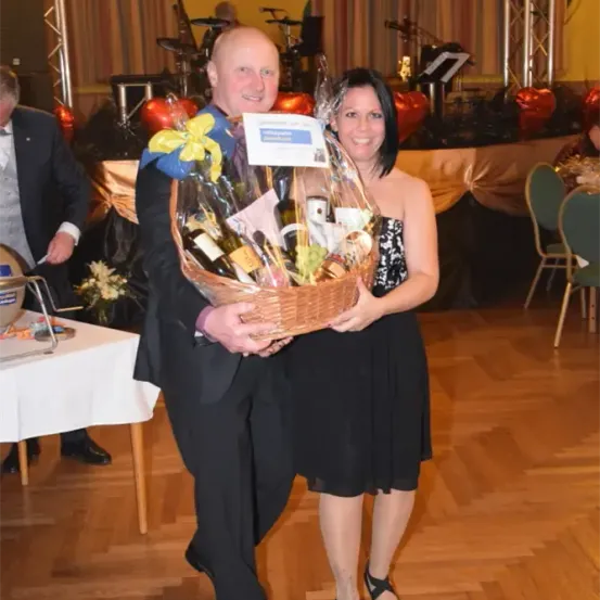 Ein Mann und eine Frau halten einen Korb mit Weinflaschen, verpackt in Plastik, bei einer formellen Veranstaltung. Dahinter steht ein Tisch mit einer weißen Tischdecke und Musikinstrumente.