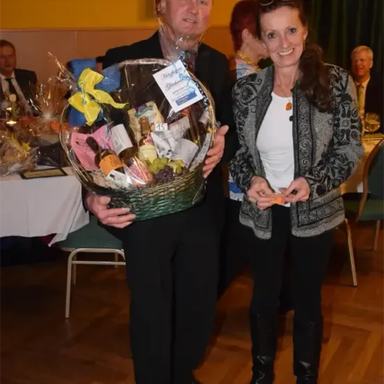 Ein Mann und eine Frau stehen lächelnd da und halten einen Geschenkkorb. Der Mann hält einen grünen Korb voller verschiedener Gegenstände, während die Frau eine kleine Karte in der Hand hält. Sie befinden sich in einem Raum mit Holzboden und Tischen mit Tischdecken.