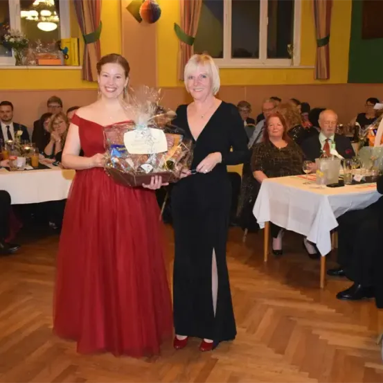 Zwei Frauen stehen in einem Saal und halten einen Korb. Dahinter sitzen mehrere Personen an Tischen, einige beobachten sie.