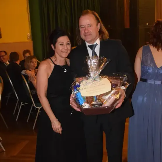 Ein Mann und eine Frau in formeller Kleidung, lächelnd, stehen zusammen und halten einen Korb mit Geschenken. Sie sind bei einer formellen Veranstaltung, mit Leuten, die in Stühlen hinter ihnen sitzen.