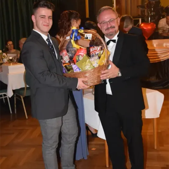 Zwei Männer stehen nebeneinander, einer hält einen Korb voller Snacks und der andere lächelt. Hinter ihnen befindet sich ein Raum mit Tischen und Stühlen, der für eine Veranstaltung dekoriert ist.