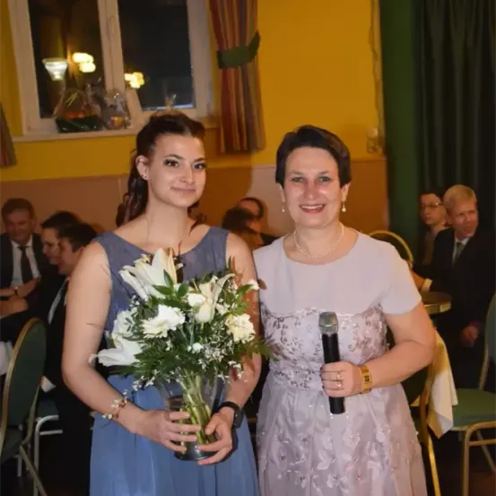 Zwei Frauen stehen lächelnd für ein Foto. Eine hält einen Blumenstrauß, die andere ein Mikrofon. Hinter ihnen ist eine gelbe Wand mit einem Fenster und Vorhängen zu sehen. Im Hintergrund sitzen mehrere Personen, darunter ein Mann mit Brille.