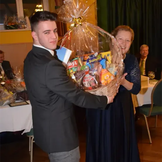 Ein junger Mann in einem Anzug hält einen Korb voller verschiedener Snacks neben einer lächelnden Frau in einem blauen Kleid. Dahinter steht ein Tisch, der mit Weingläsern und anderen Gegenständen gedeckt ist. Ein Mann in Anzug und Krawatte sitzt im Hintergrund.