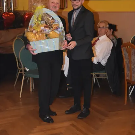 Zwei Männer in Anzughose stehen in einem Raum, einer hält einen Geschenkkorb und der andere lächelt. Hinter ihnen beobachtet ein sitzender Mann in einem weißen Hemd und roter Krawatte. Grüne Stühle, ein Holzboden und eine gelbe Wand mit Dekorationen sind sichtbar.