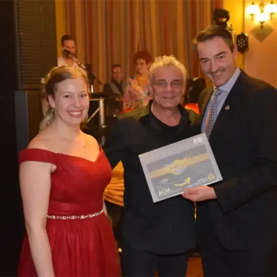 Drei Menschen stehen lächelnd und halten ein gerahmtes Dokument. Eine Frau in Rot trägt eine Halskette, und ein Mann in einem schwarzen Anzug lächelt mit einer Anstecknadel. Ein Mann in einem schwarzen Hemd steht hinter ihnen.