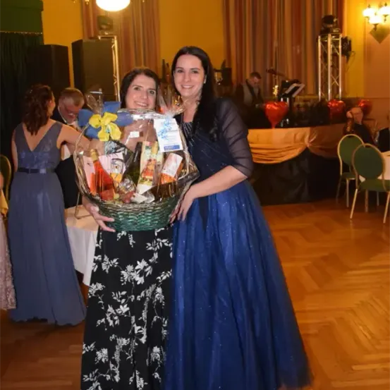 Zwei Frauen in Abendkleidern stehen zusammen und halten einen Geschenkkorb mit verschiedenen Gegenständen. Hinter ihnen ist ein Raum mit Stühlen und einer Bühneneinrichtung zu sehen.