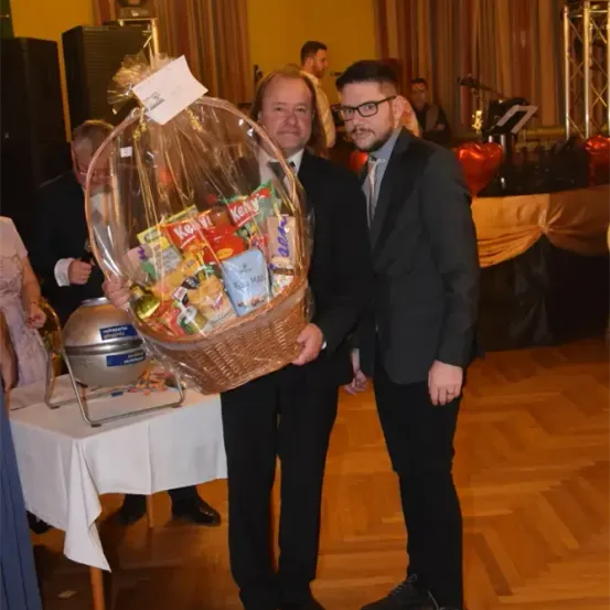 Zwei Männer in Anzügen halten einen Korb mit Snacks bei einer Veranstaltung. Hinter ihnen tummeln sich Leute und ein Tisch mit einem Kuchen ist zu sehen.