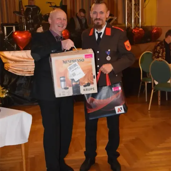 Zwei Männer präsentieren ein Geschenk, wobei einer eine Box einer Nespresso-Kaffeemaschine hält und der andere eine Tasche trägt. Hinter ihnen ist ein dekorierter Hintergrund mit einem Tisch und Stühlen.