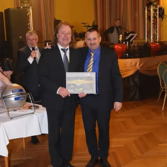 Zwei Männer in Anzügen halten eine Preisplakette während einer Veranstaltung. Ein dritter Mann hält ein Mikrofon und spricht. Dahinter spielt ein weiterer Mann ein Instrument.