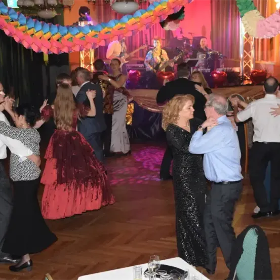 Eine lebhafte Tanzfläche mit farbenfrohen Dekorationen, an der eine Band auf der Bühne mit verschiedenen Instrumenten spielt. Die Tänzer, in formeller Kleidung gekleidet, amüsieren sich.