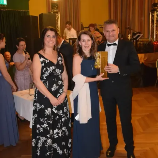 Drei Personen stehen in einem formellen Rahmen zusammen, eine Frau hält eine Trophäe, eine andere lächelt und ein Mann mit Fliege schaut in die Kamera. Hinter ihnen unterhalten sich weitere Personen bei einer Veranstaltung.