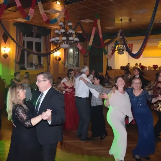 Eine Gruppe von Menschen tanzt in einem dekorierten Raum. Einige tragen Abendgarderobe, andere Cocktailkleider. Der Raum hat eine Kronleuchter und bunte Dekorationen. Eine Person steht in der Nähe eines Fensters.